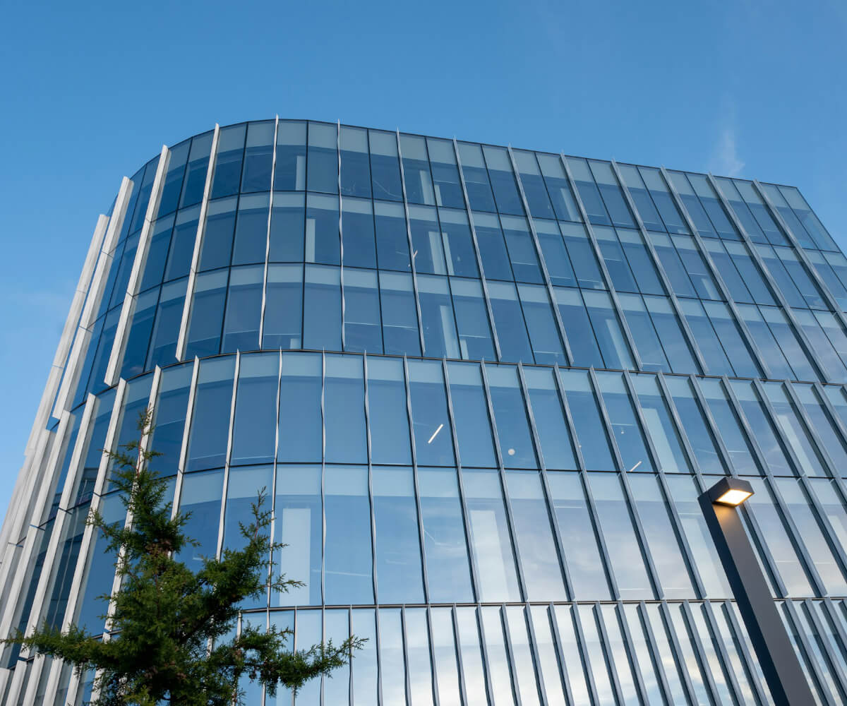Glass office building with reflective windows against a clear blue sky, with a tree and a streetlight in the foreground.