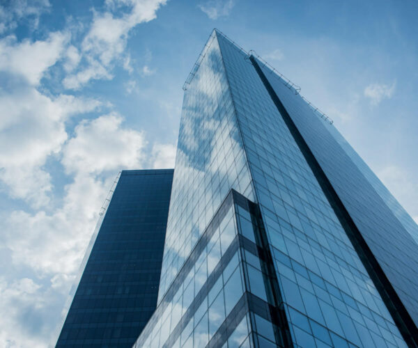 Two modern glass skyscrapers against a cloudy sky, with reflections visible on their surfaces.