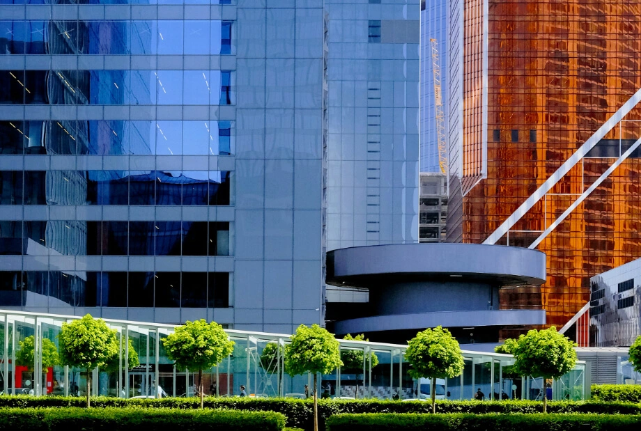 Modern cityscape with glass skyscrapers, reflective surfaces, and a row of neatly trimmed green trees in the foreground.