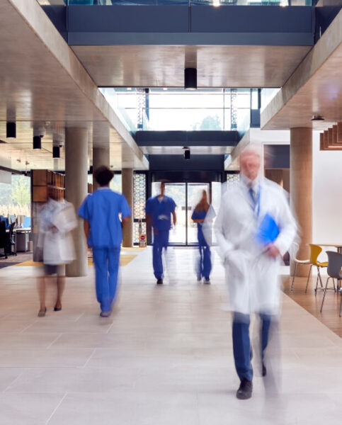 Medical professionals in white coats and blue scrubs walk through a modern hospital corridor with glass walls and open spaces.