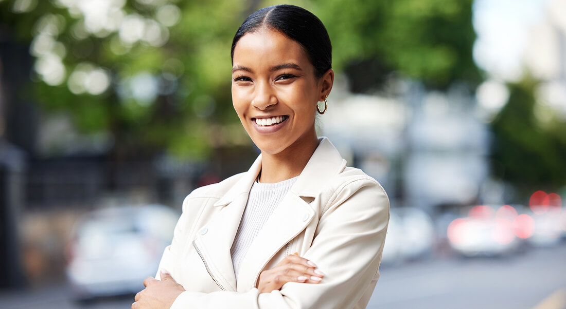 A woman with dark hair pulled back, wearing a light-colored jacket and hoop earrings, stands outdoors with arms crossed, smiling. Blurred street and trees in the background.