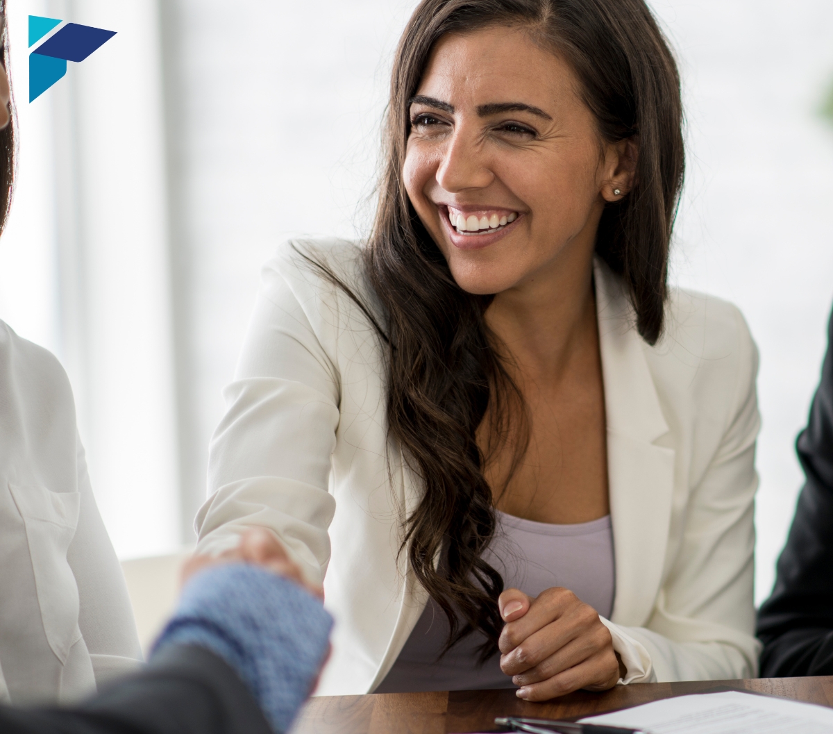 A smiling woman in a white blazer shakes hands with another person in a business setting.