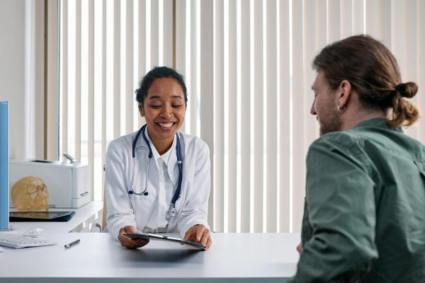 A doctor in a white coat talks with a patient across a desk, holding a clipboard. Vertical blinds cover the window in the background.