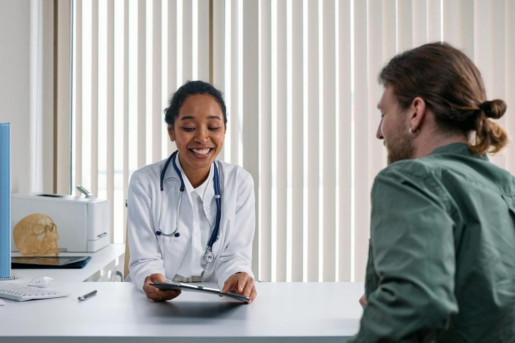 A doctor in a white coat reviews a clipboard with a patient seated across the desk in a medical office with vertical blinds.