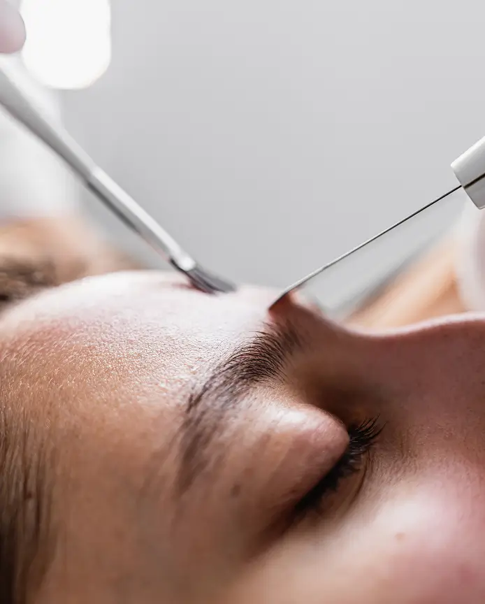 Close-up of a person's face receiving a cosmetic eyebrow or skin treatment with metal tools near the eyebrow area.