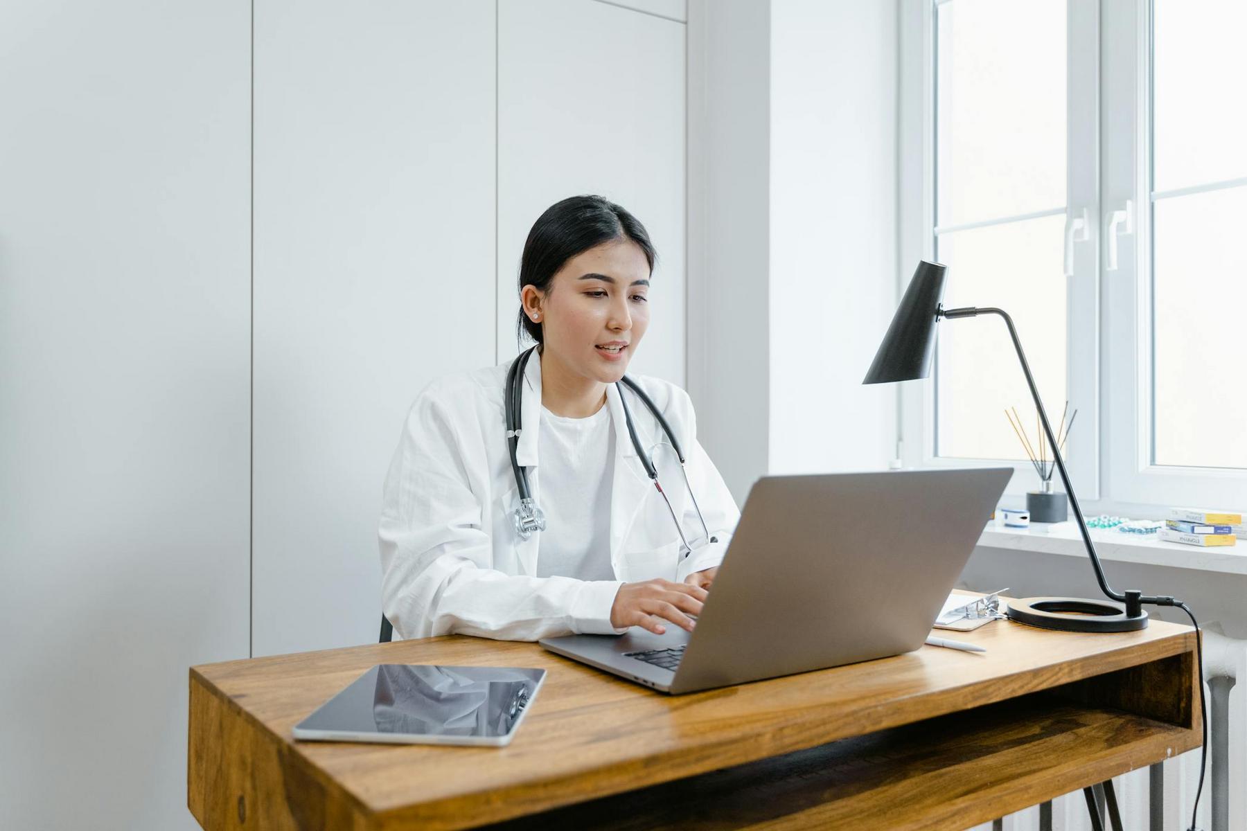 A doctor wearing a white coat and stethoscope sits at a desk, working on a laptop. A tablet, lamp, and other items are on the wooden desk, with a window in the background.