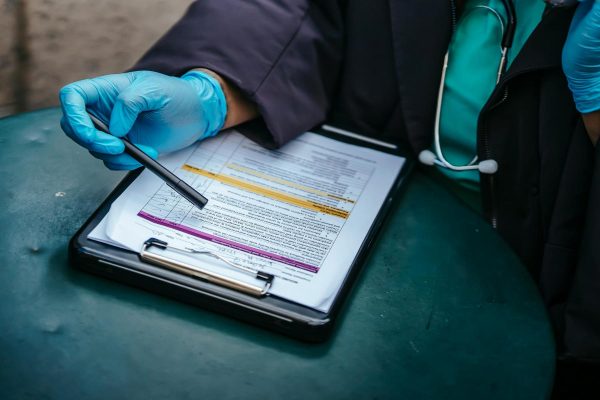 Person wearing blue medical gloves points to a form on a clipboard with a pen; a stethoscope is visible around their neck.