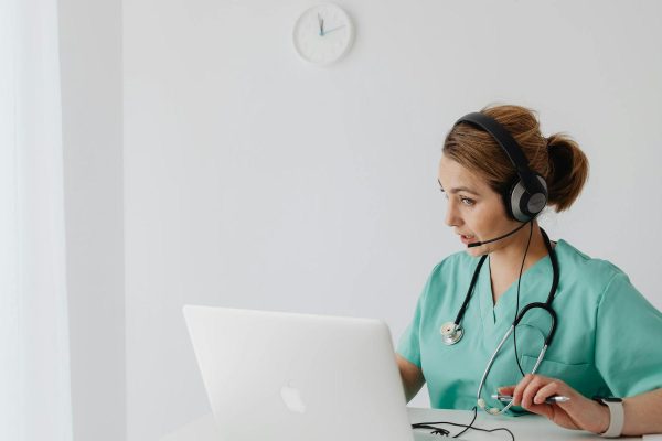 A healthcare professional in scrubs and a stethoscope wears a headset while working at a laptop in a bright, minimalist room with a clock on the wall.